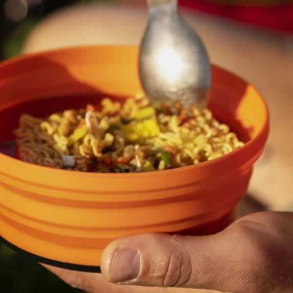 A spoon hovers over and orange bowl filled with noodles.
