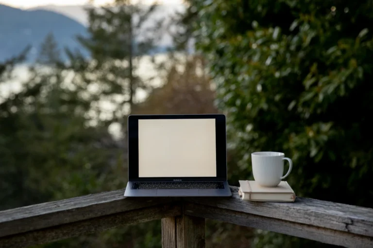 A computer and coffe cup sit on a porch railing.