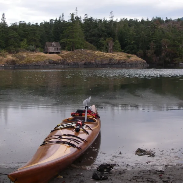 A cedar strip kayak sits on a beach across the bay from an old house.