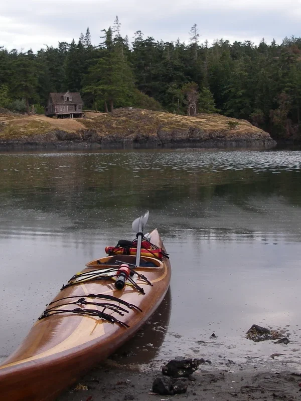 A cedar strip kayak sits on a beach across the bay from an old house.