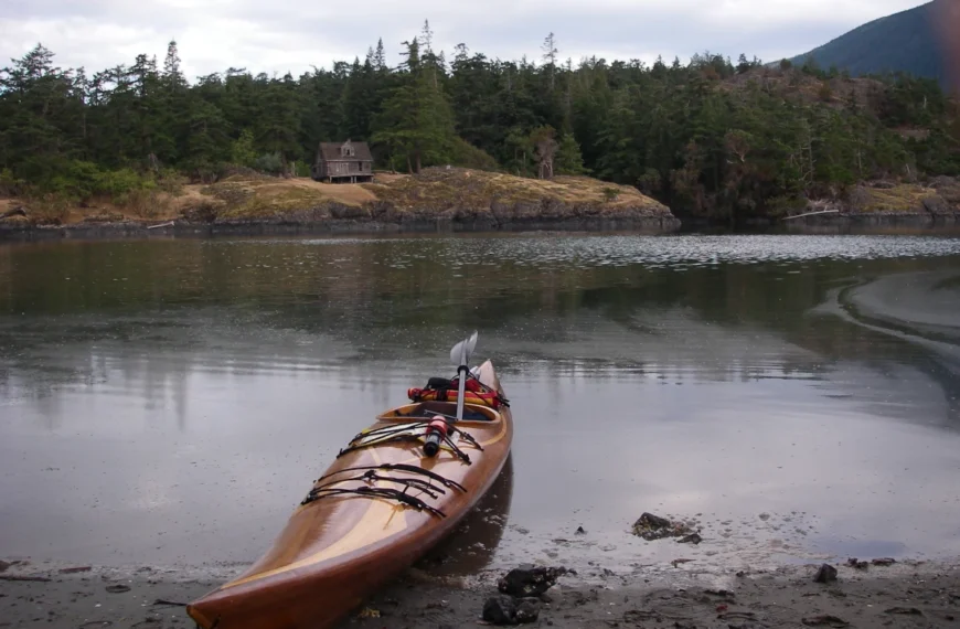 A cedar strip kayak sits on a beach across the bay from an old house.