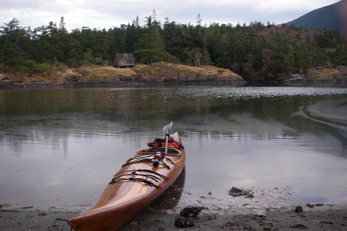 A cedar strip kayak sits on a beach across the bay from an old house.