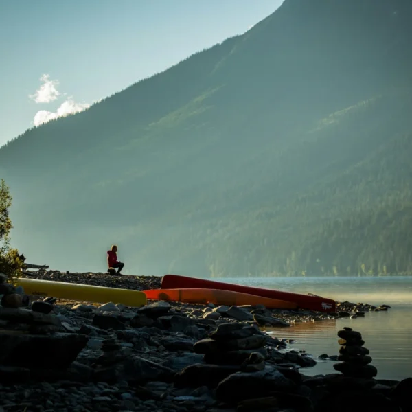 A man sits near canoes on a rocky lakeshore.