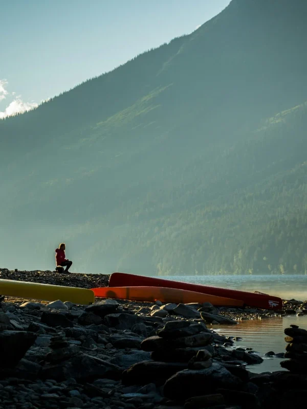 A man sits near canoes on a rocky lakeshore.