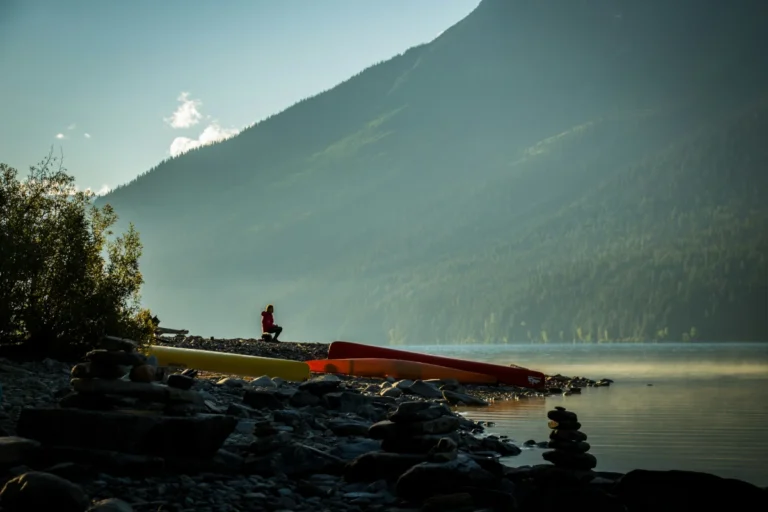 A man sits near canoes on a rocky lakeshore.