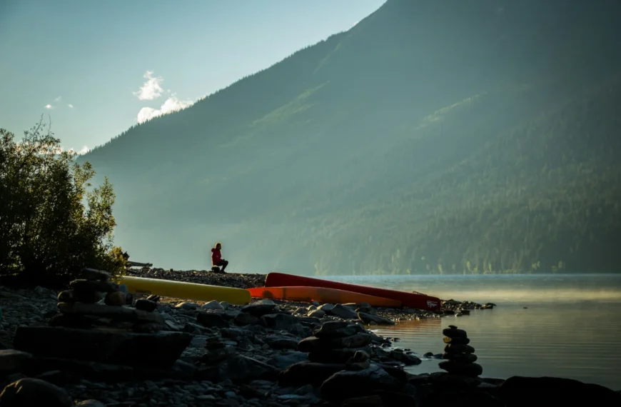 A man sits near canoes on a rocky lakeshore.