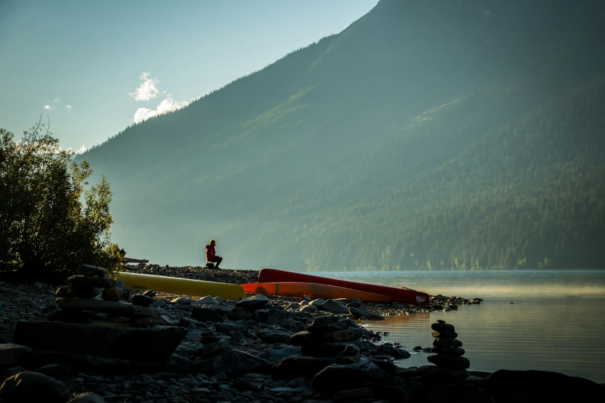 A man sits near canoes on a rocky lakeshore.
